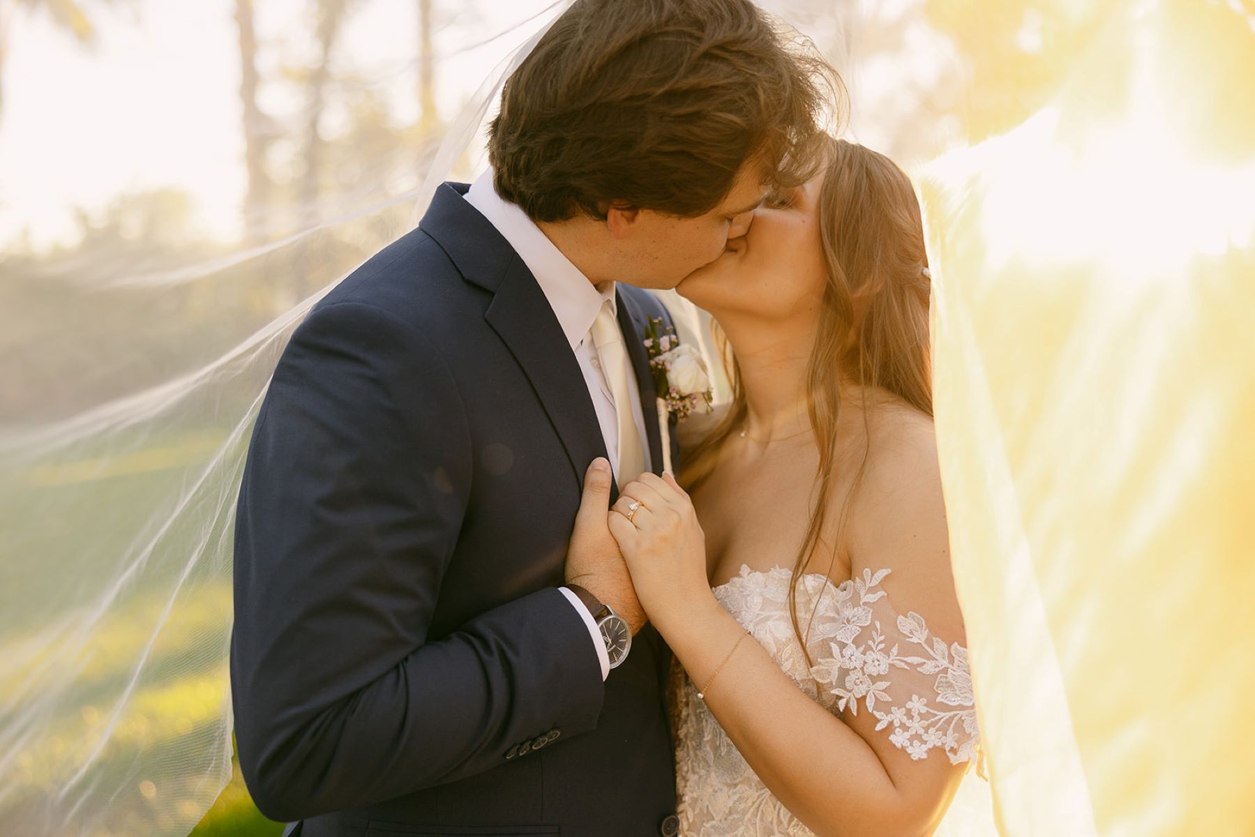 A couple kissing underneath of the bride's wedding veil and holding hands the bride is wearing a white wedding dress and the groom is wearing a tuxedo 