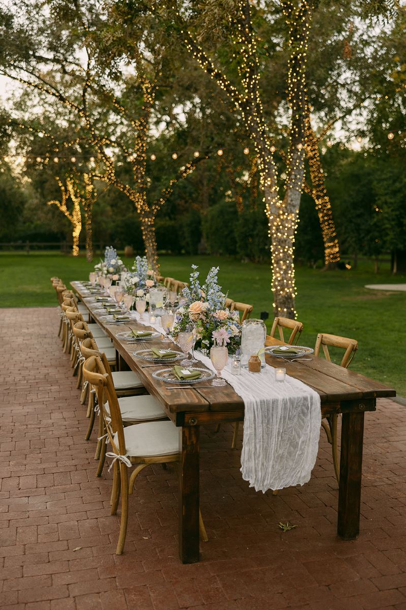 A long wooden table with wooden chair decorated with a white cloth and flowers in vases, glasses, plates, and silverware in the background is a lawn with trees that are wrapped in yellow lights 