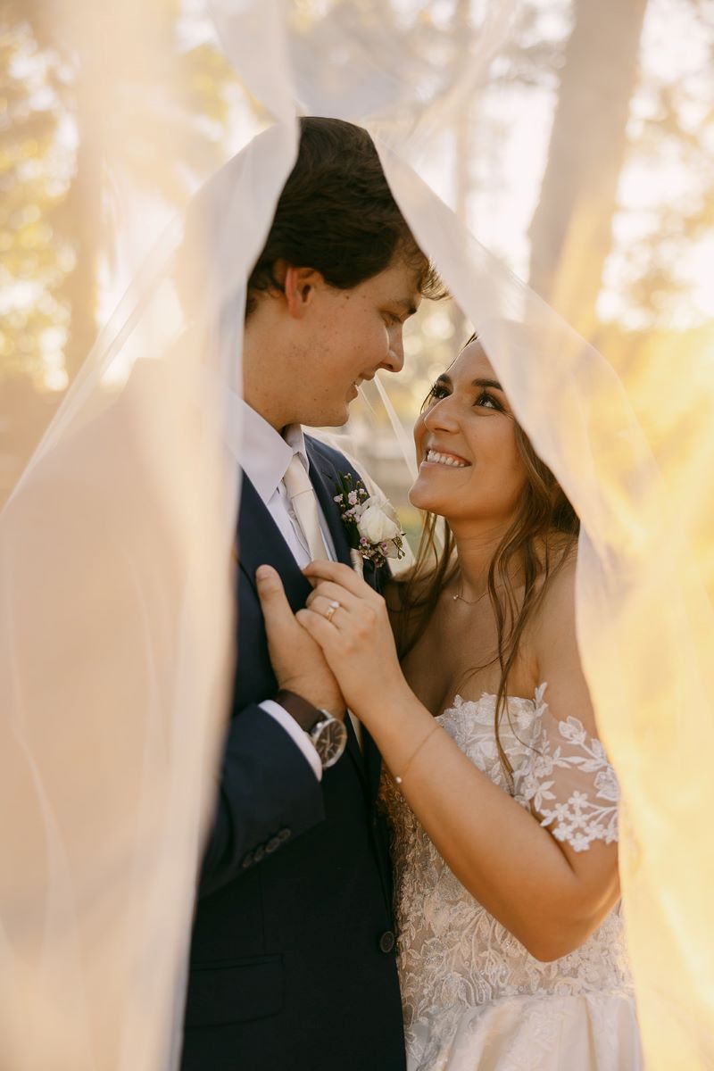 A couple smiling at each other underneath of the bride's wedding veil and holding hands and looking into each other's eyes the bride is wearing a white wedding dress and the groom is wearing a tuxedo 