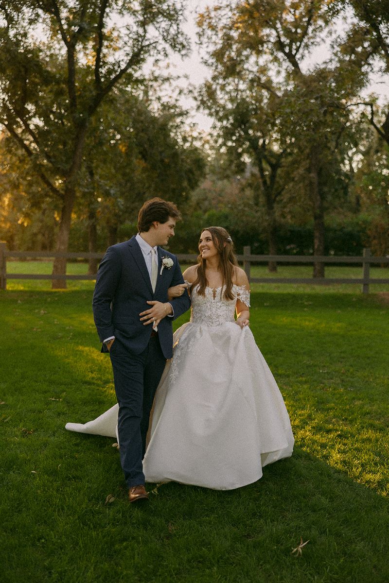 A couple walking through a green field together looking at each other the bride is holding the groom's arm the bride is wearing a white wedding dress and the groom is wearing a tuxedo 