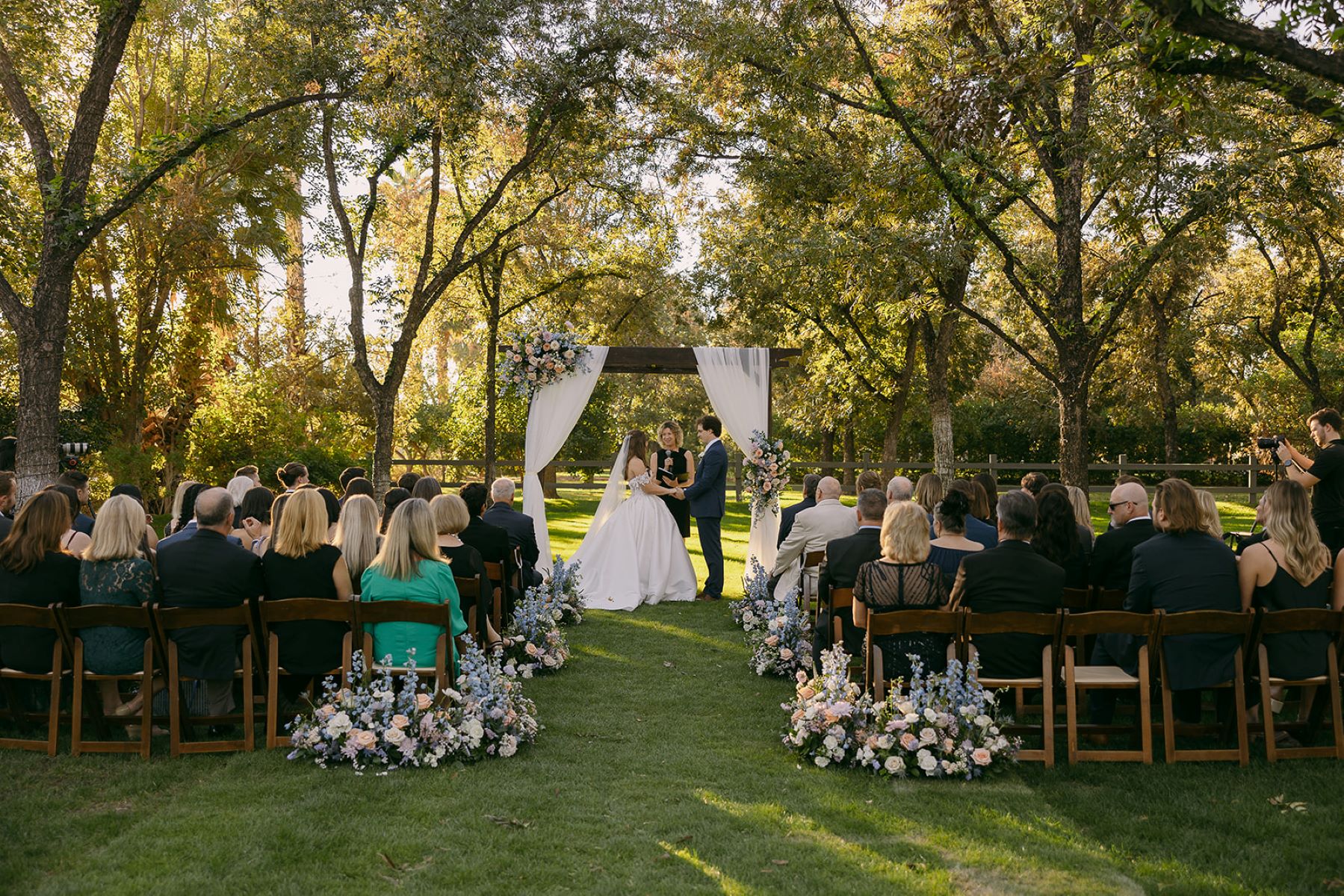 A couple at their wedding ceremony with their officiant with their guests sitting on wooden chairs in front of them watching the bride is wearing a white wedding dress and veil and the groom is wearing a tuxedo and they are holding hands 