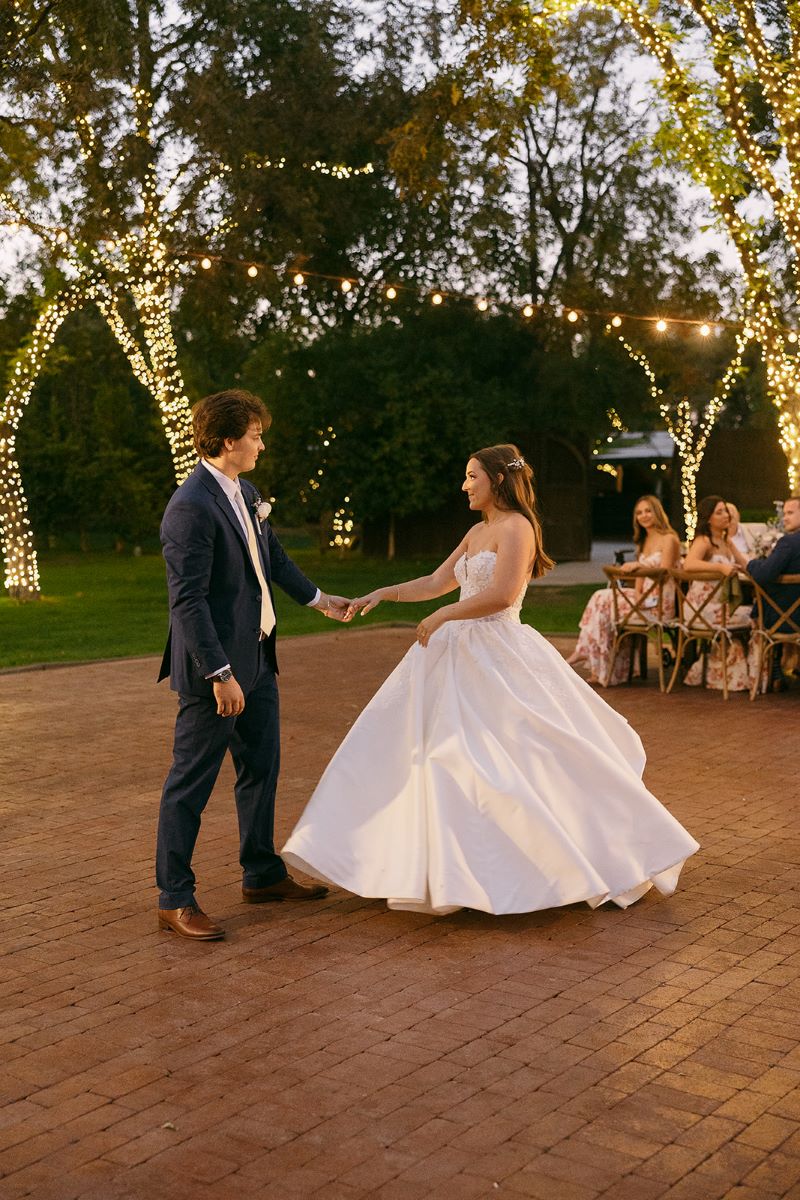 A couple at their wedding reception dancing on a brick dance floor as their guests watch behind them the trees are wrapped in yellow lights the bride is wearing a white wedding dress the groom is wearing a tuxedo 