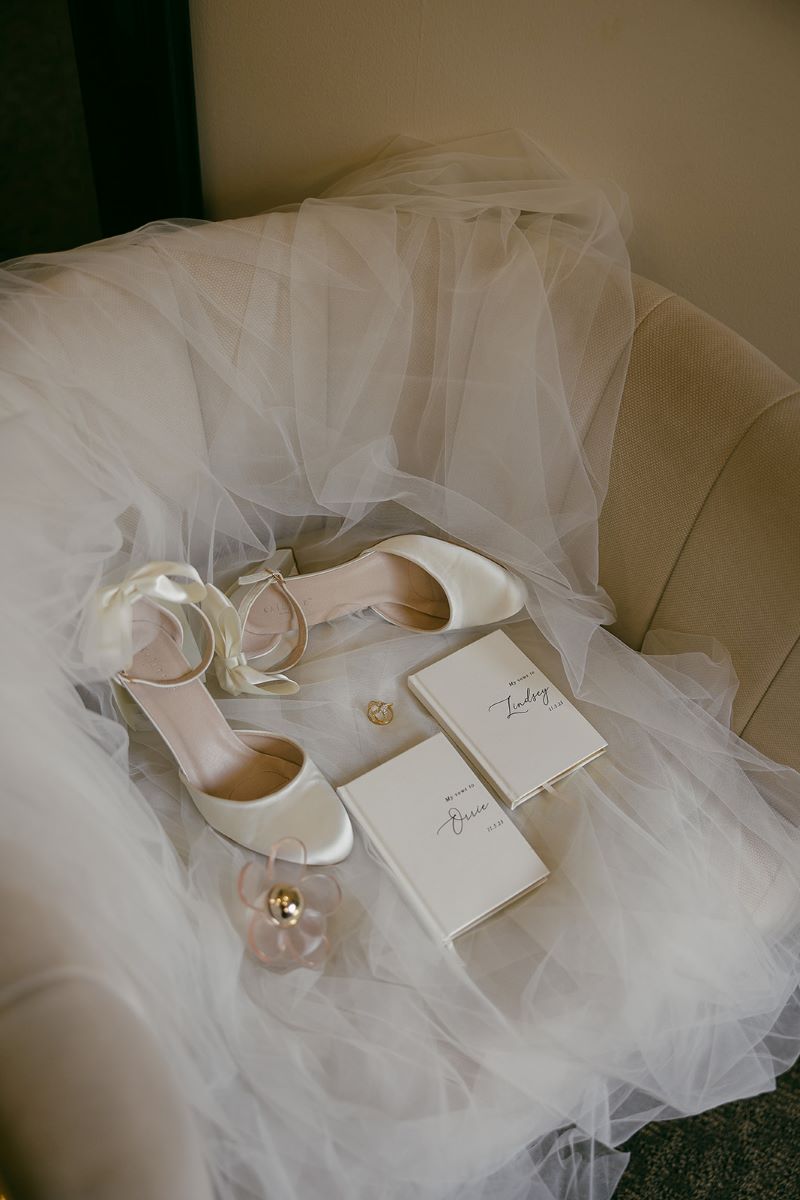 A white wedding veil on a chair and on top of the veil is a pair of white heels, vow books, a perfume, and a wedding ring