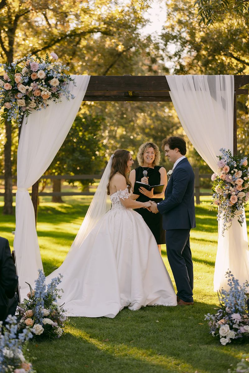 A couple at their wedding ceremony with their officiant the couple is holding hands and laughing the bride is wearing a white wedding dress and veil and the groom is wearing a tuxedo 