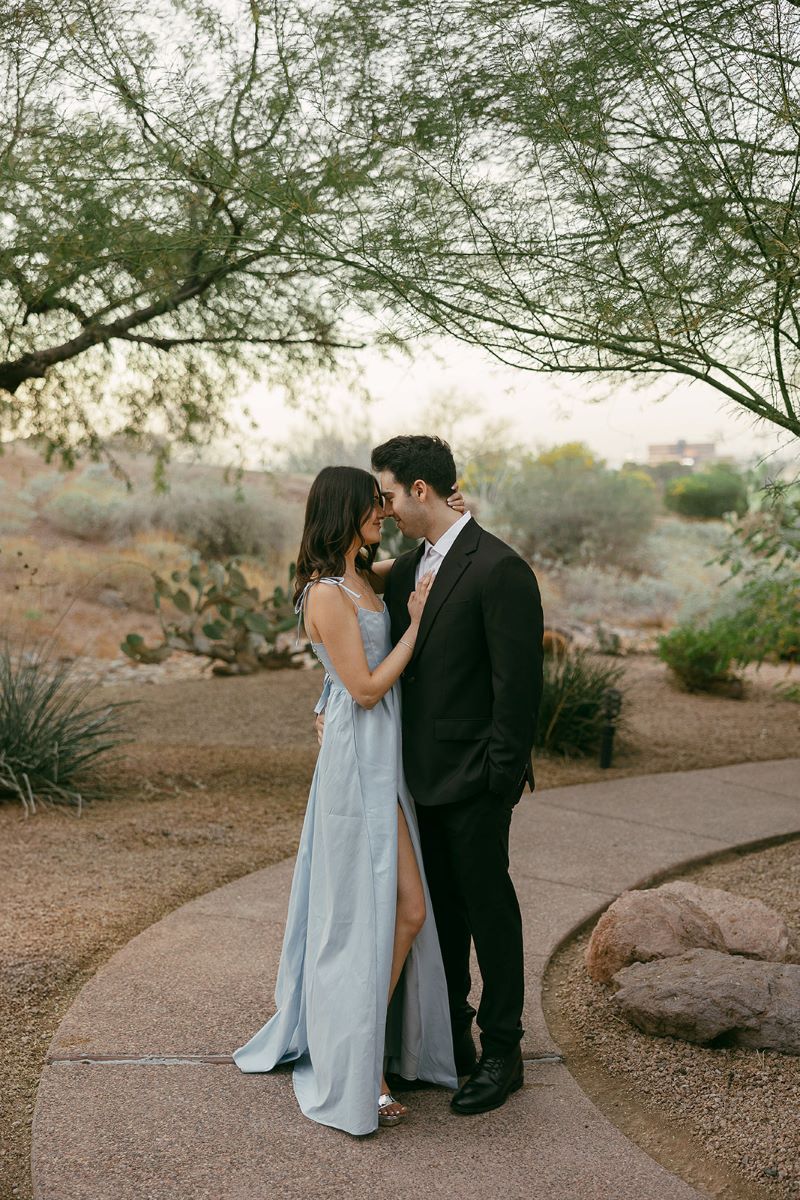 A couple standing on a sidewalk togehter looking into each other's eyes and smiling the woman is wearing a blue dress and has her hand on her partner's chest and her partner is wearing a black suit 