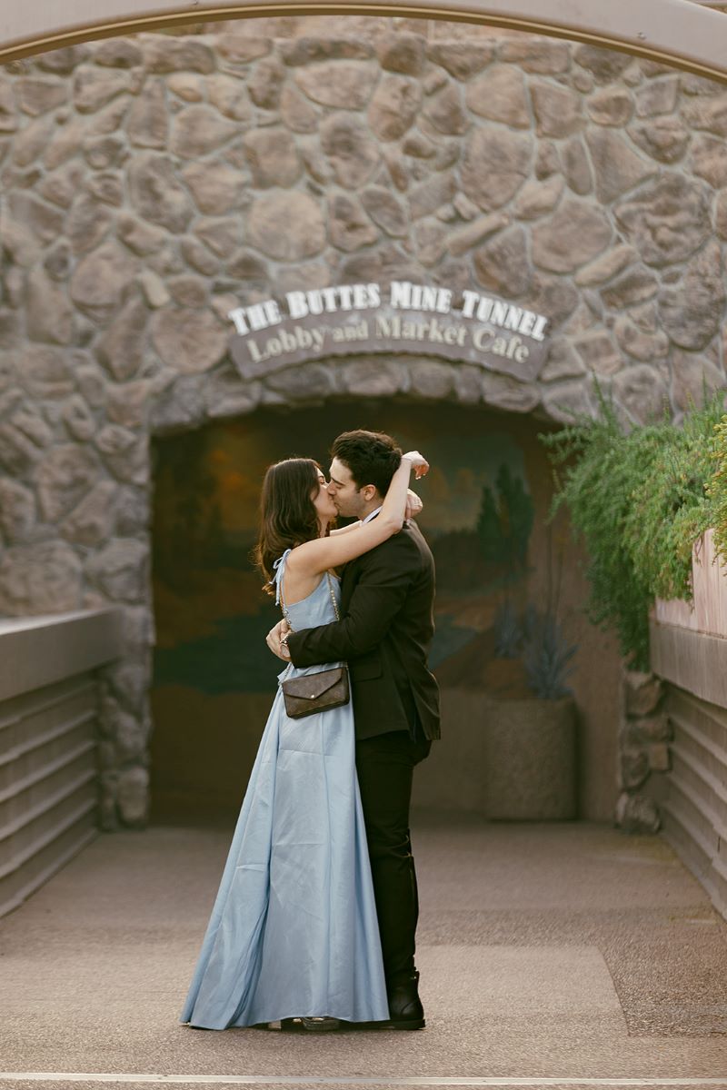 a woman in a blue dress wiht a black purse has her arms around her partner's shoulders and is kissing him her partner is wearing a black suit