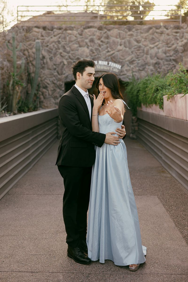 A woman showing off her engagement right she is wearing a blue dress and has her nails down her partner is smiling at her and wearing a black suit and has his arms around her