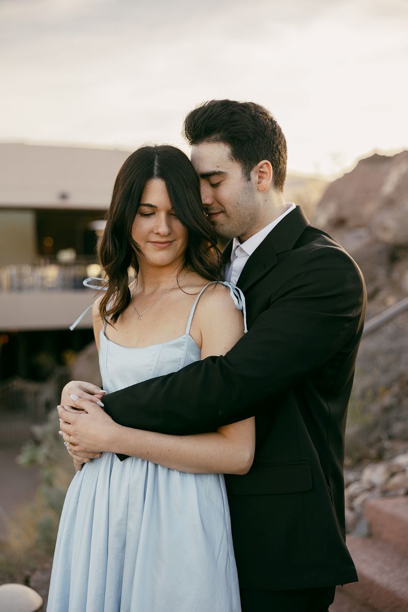 A couple together the man is wearing a black suit with his arms around his partner's waist and is pressing his nose against her hair the woman is wearing a blue dress 