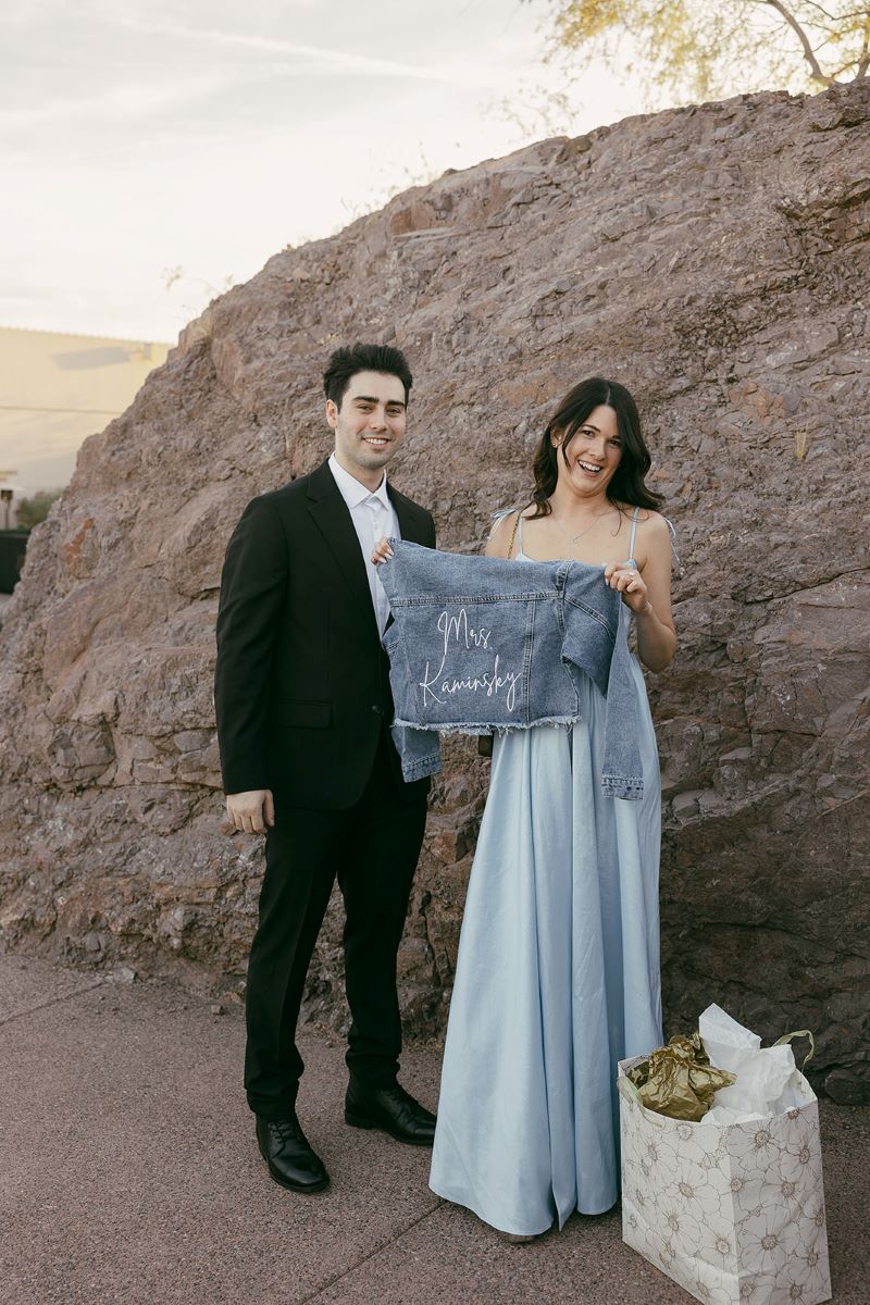 A woman holding up a blue jean jacket that has her partner's last name on in and smiling her partner is standing next to her and smiling 