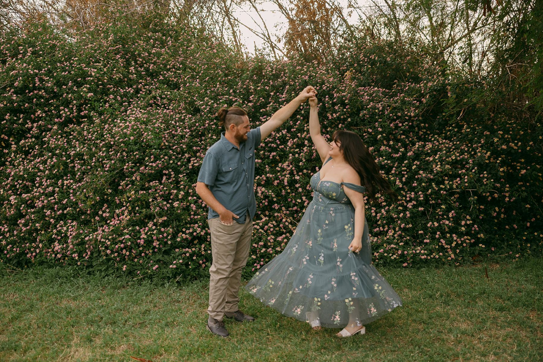 a couple dancing in fornt of a bush filled with pink flowers the woman is wearing a blue dress with flowers the man is wearing a blue button up and tan pants and is spinning his partner and smiling at her 