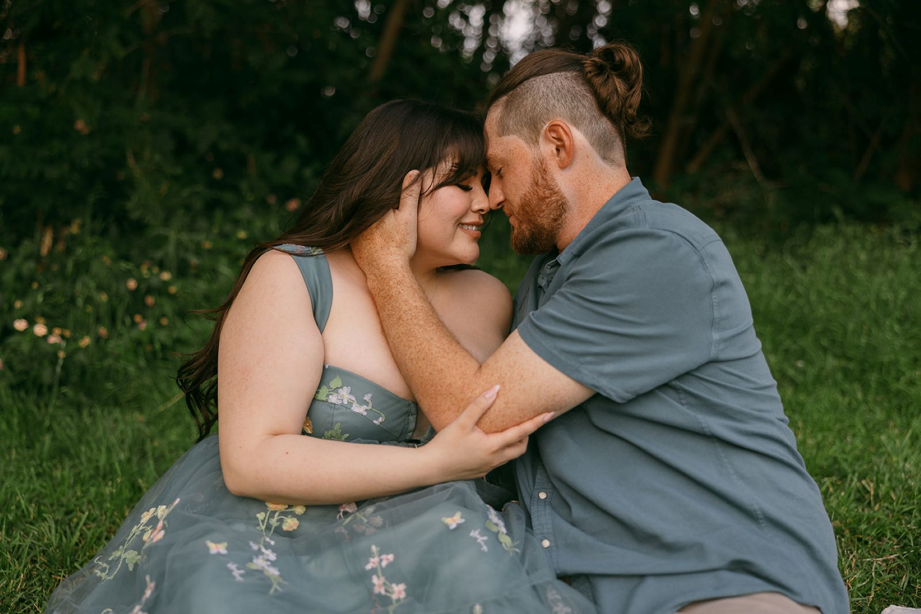 a man and woman sitting on a picnic blanket with their heads pressed together the man has his hand on the side of the woman's face the woman is touching the man's elbow and they both have their eyes closed