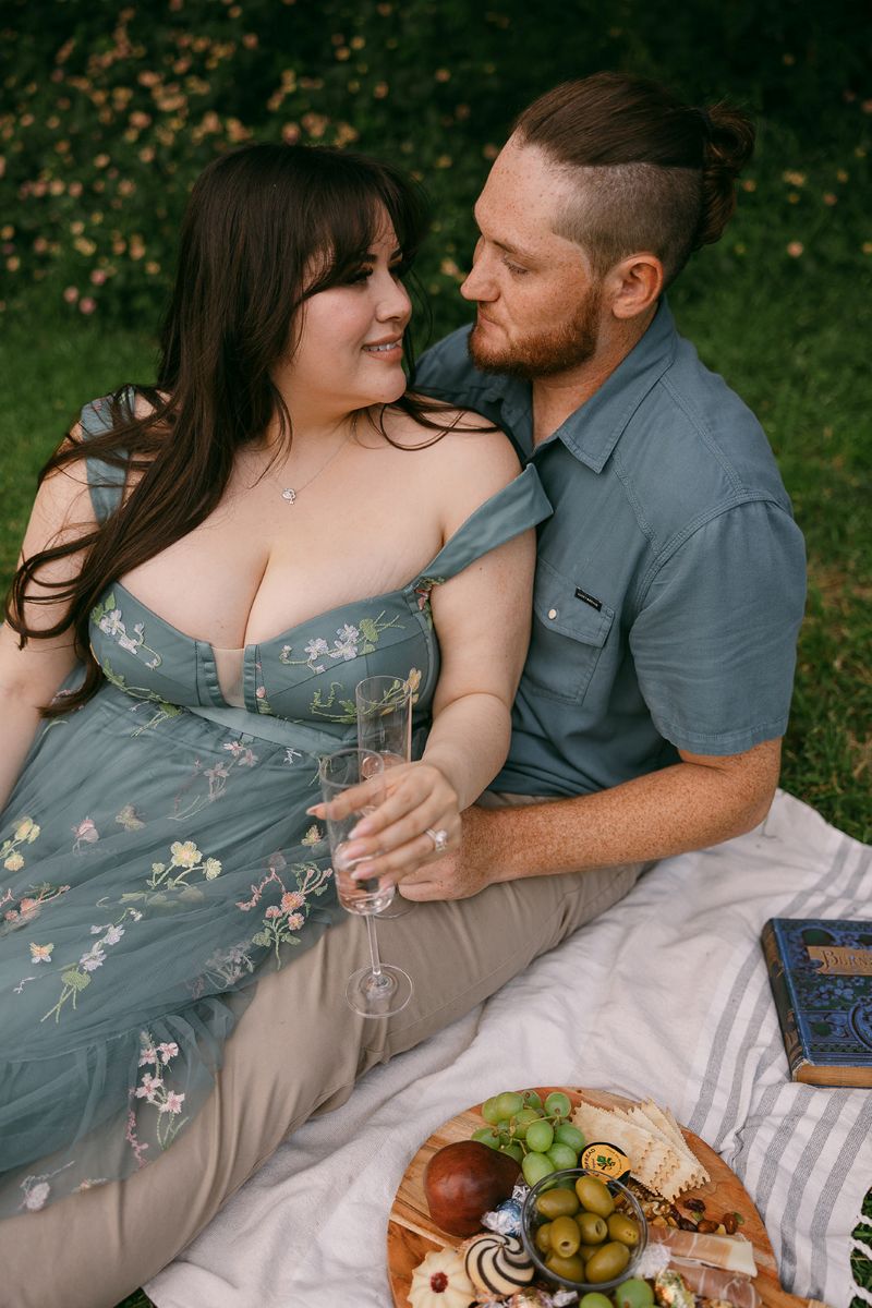 a man and woman sitting on a picnic blanket holding champagne glasses the woman is leaning back into the man and on the picnic blanket is a book and snacks 