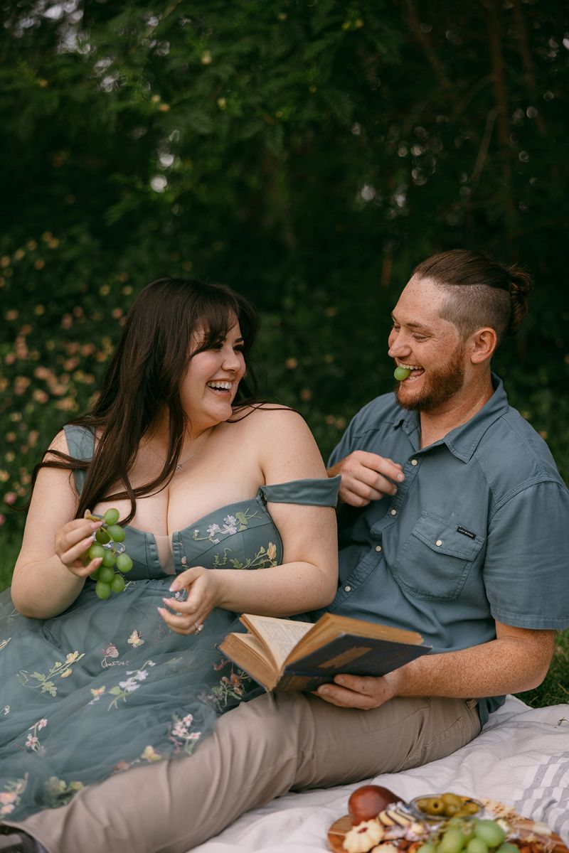 a man and woman sitting on a blanket eating grapes they are both laughing with each other the man is also holding a book and there are snacks on the blanket as well 