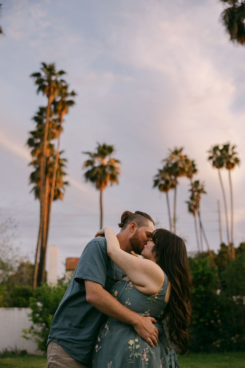 a man and woman kissing each other underneath palm tress the woman has her arms around the man's shoulders the man has his arms around her waist the woamn is wearing a blue dress with flowers and the man is wearing a blue button up 