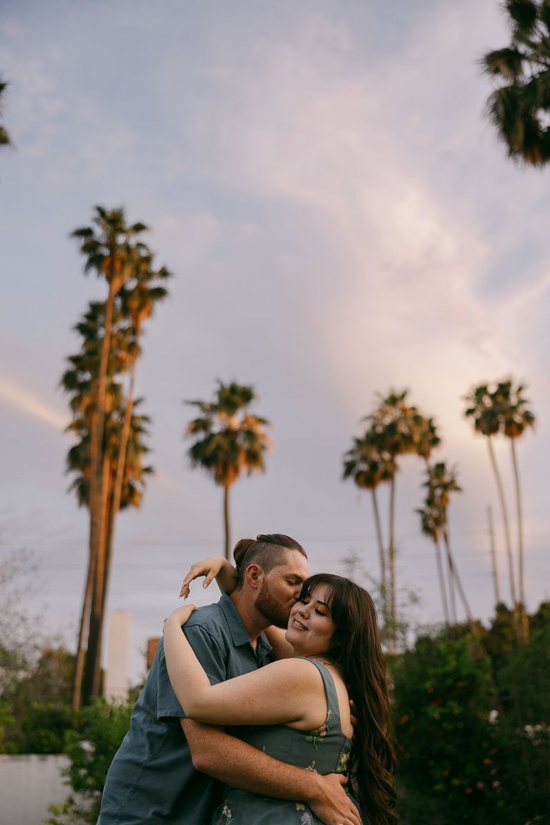 a man kissing the side of a woman's cheek and they are standing underneath palm trees the woman is hugging the man around the shoulders and the man is hugging the woman around her waist 