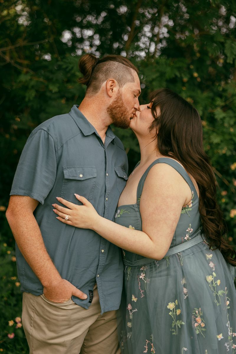 a couple kissing one another the woman has her engagement ring on and is touching the man side and the man has his hand in his pocket the man is wearing a blue button up and the woman is wearing a blue floral dress 