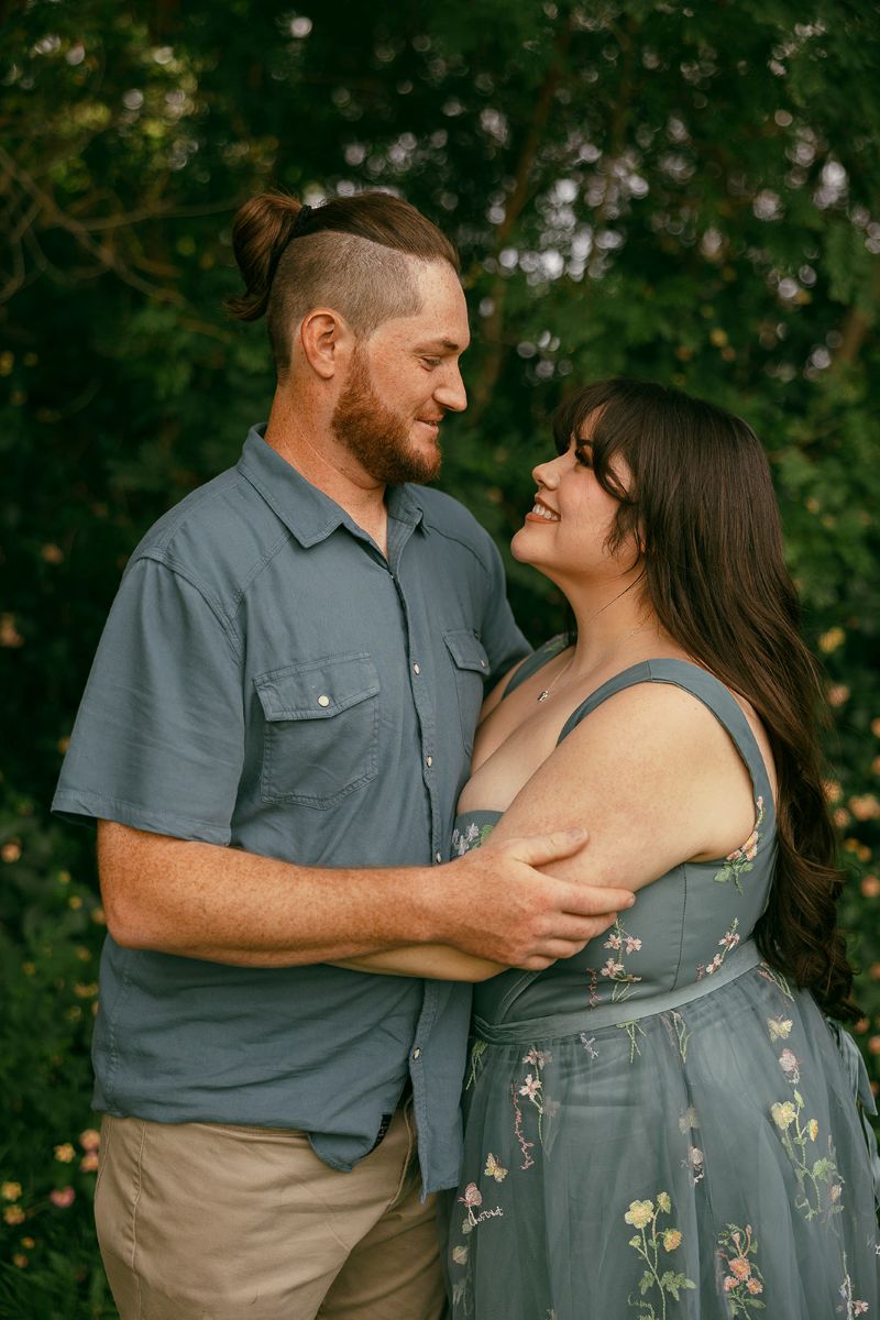 a man and woman embracing and smiling at each other the woman is wearing a blue dress with flwoers the man is wearing a blue button up 