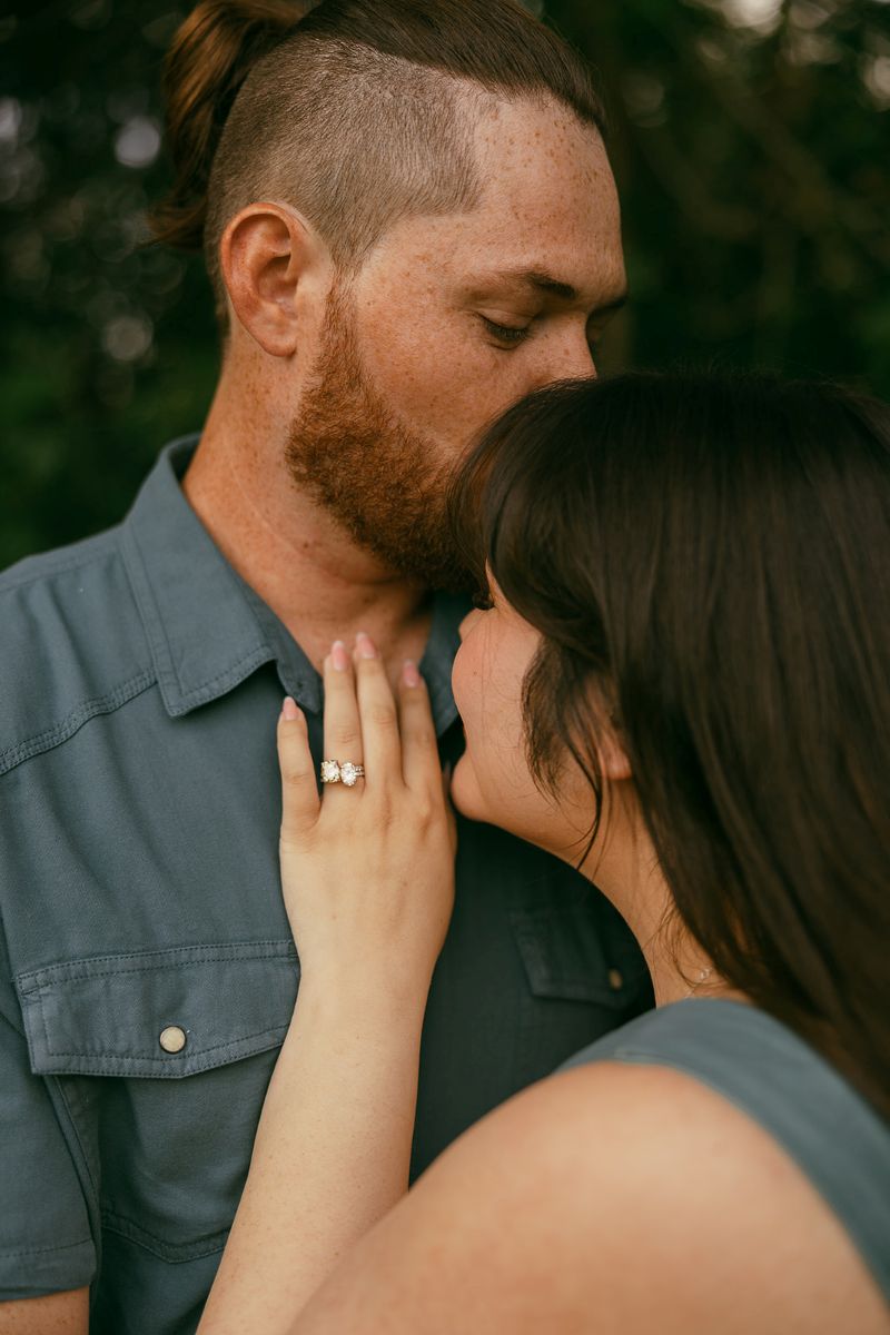 a man is kissing his partner on the forehead and the woman is smiling and has her hand on his chest and she is wearing her engagement ring 