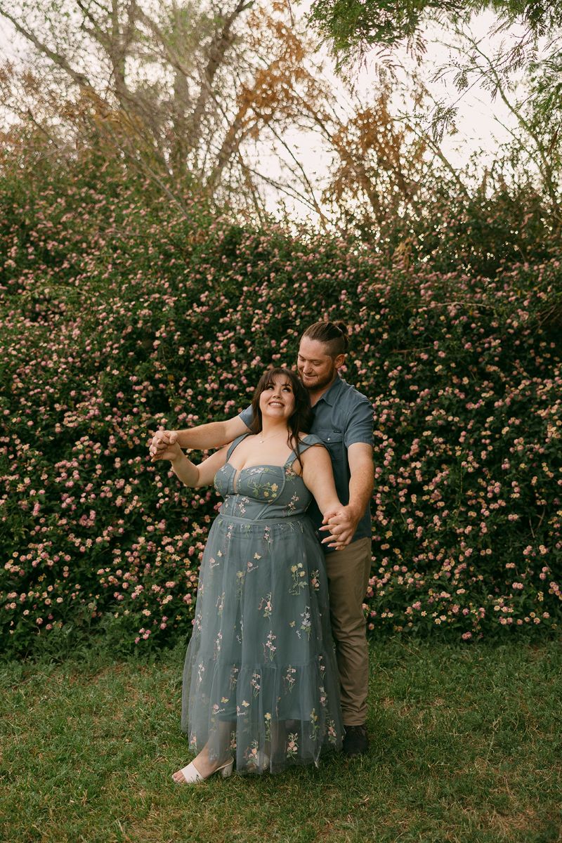 a couple in front of a bush with pink flowers the man is behind his partner and holding out her arms to this side the woman is leaning back into him and smiling she is wearing a blue dress with flowers the man is wearing a blue shirt with tan pants 