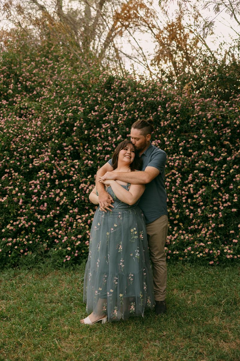 a man and woman in front of a large bush with pink flwoers the man is hugging the woman from behind and kissing the side of her head the woman is leaning back into the man and smiling 