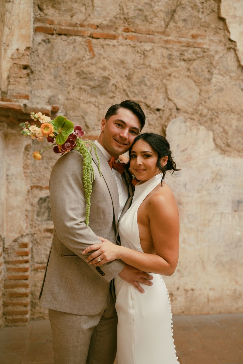 a copule embracing and smiling inside of an old chapel the woman is wearing a white wedding dress and holding flowers the man is wearing a gray suit