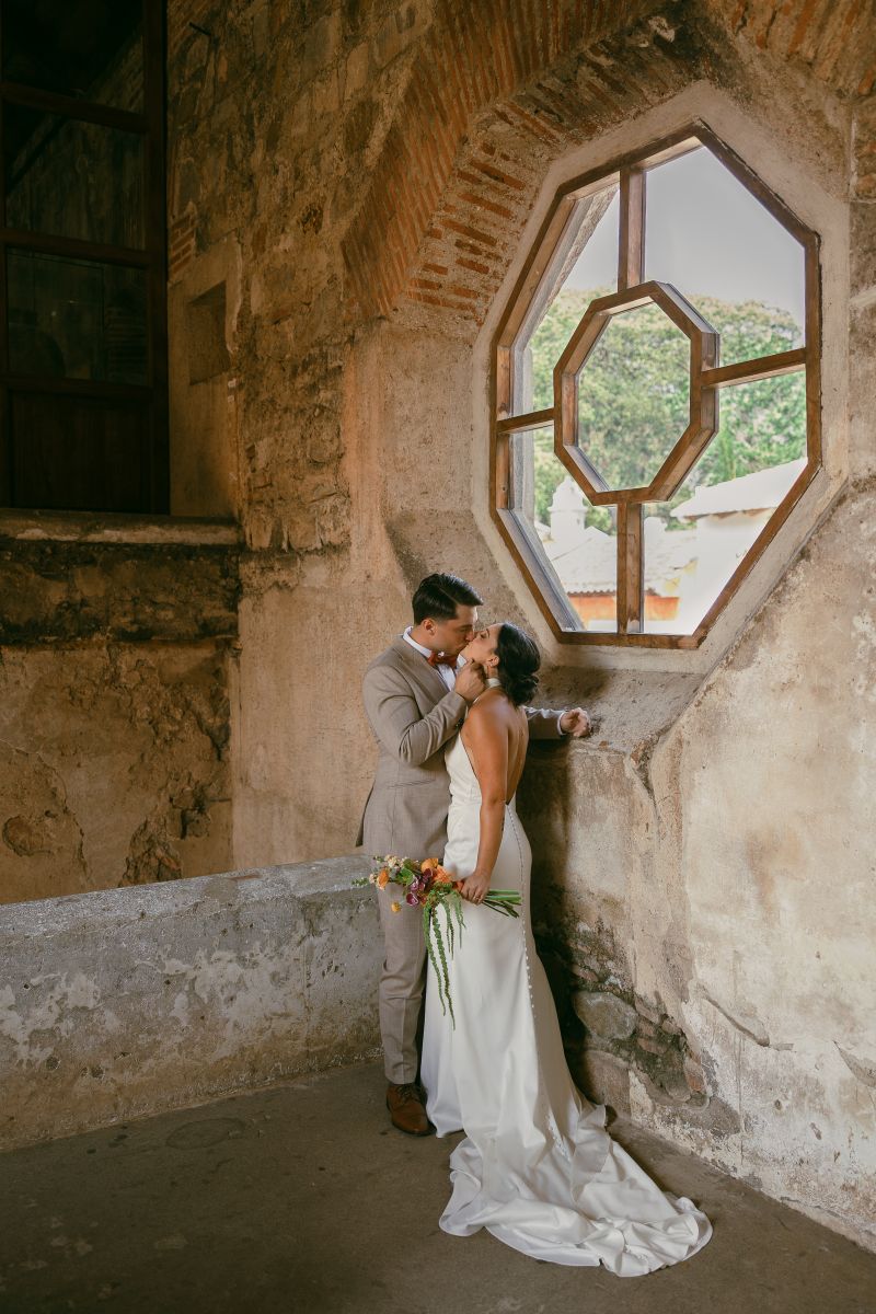 a couple kissing inside convento capuchinas in antigua guatemala the woman is wearing a wedding wedding dress and holding flwoers the man is wearing a gray suit