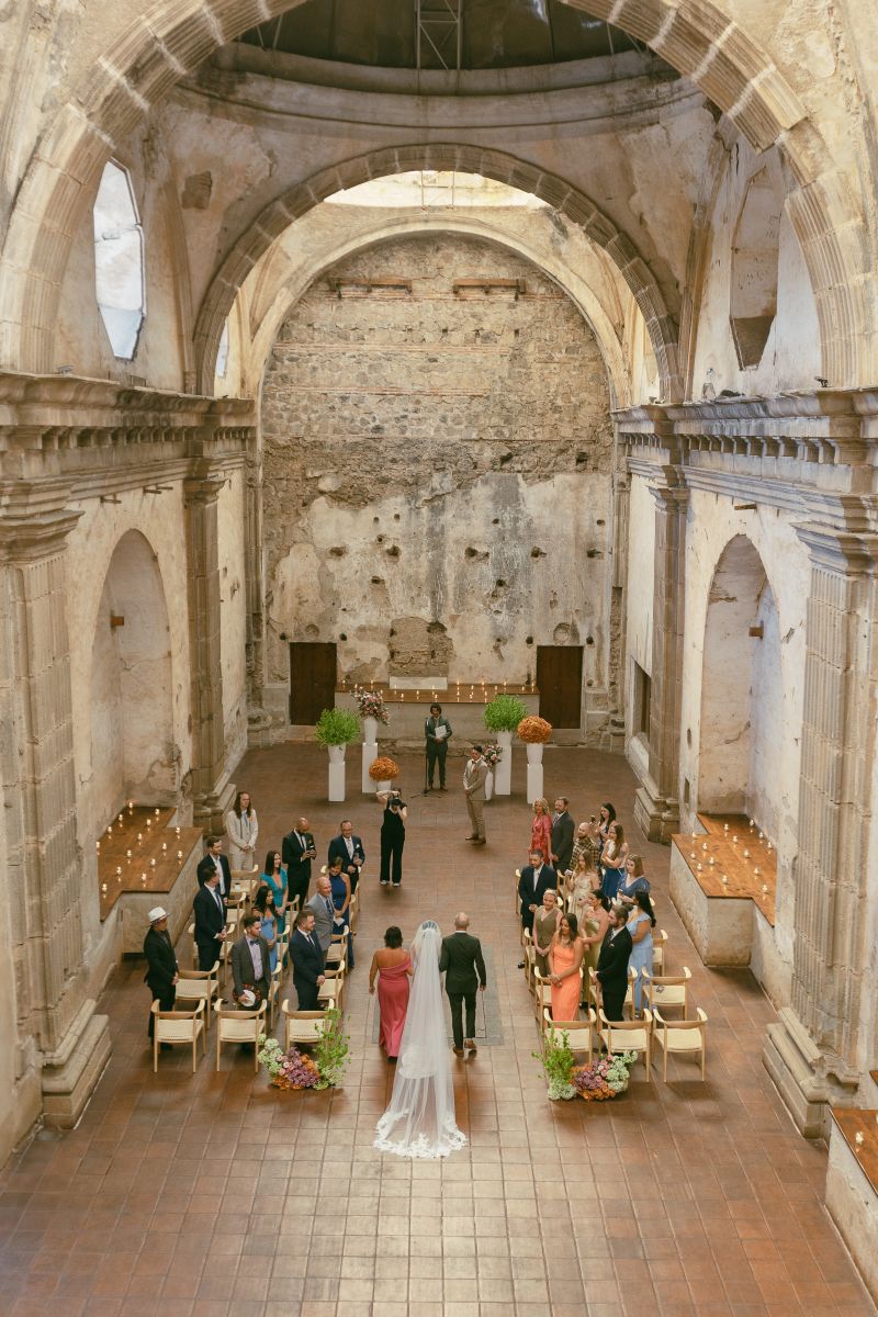 a mother and father walking their daughter down the aisle to her wedding ceremony in convento capuchinas chapel in antigua guatemala