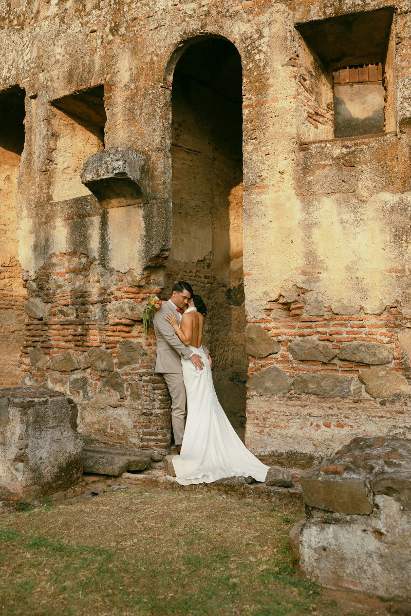 a man embracing his partner in historic ruins in antigua Guatemala the woman is wearing a white wedding dress and holding flowers the man is wearing a tan suit